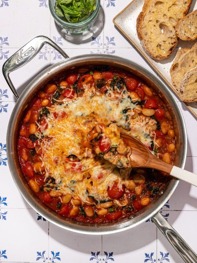 skillet with cheesy white beans in pizza sauce and wooden spoon beside sliced bread and fresh basil