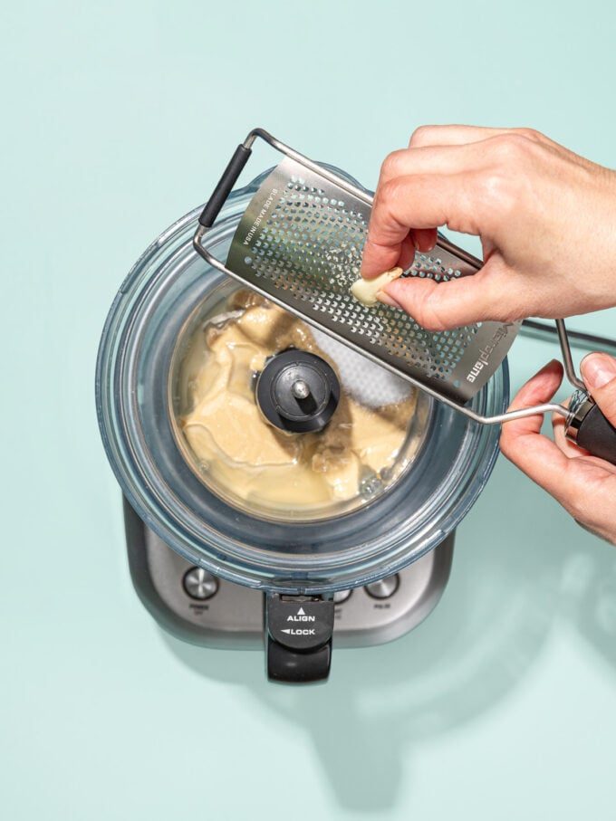 hand microplaning garlic into food processor with tahini
