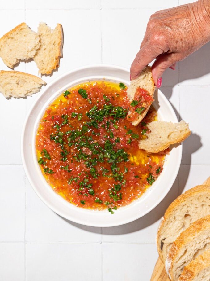 hand dipping bread into olive oil and tomato bread dip on white plate with slices of bread scattered around it.