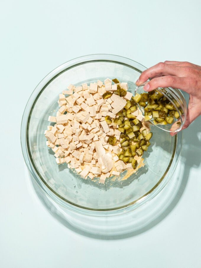 hand pouring chopped pickles into glass bowl with diced tofu and dressing