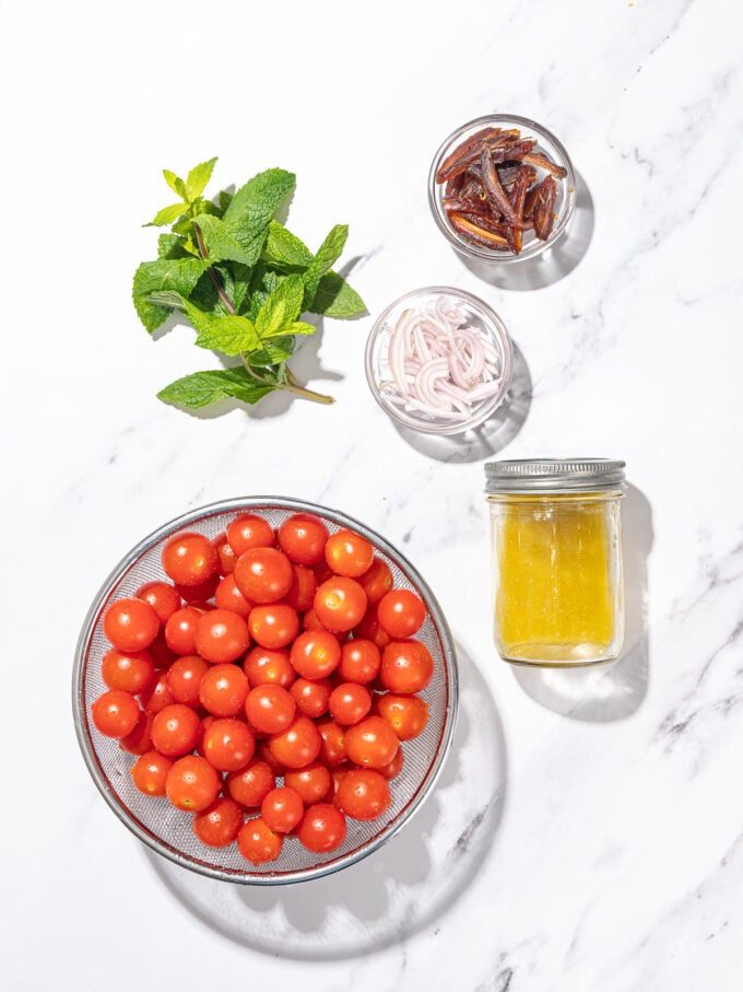 glass bowl of cherry tomatoes, beside jar of garlic dressing, mint, sliced dates and shallots